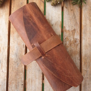 May include: A brown leather roll-up case with a strap closure. The case is made of distressed leather and has a rustic look.