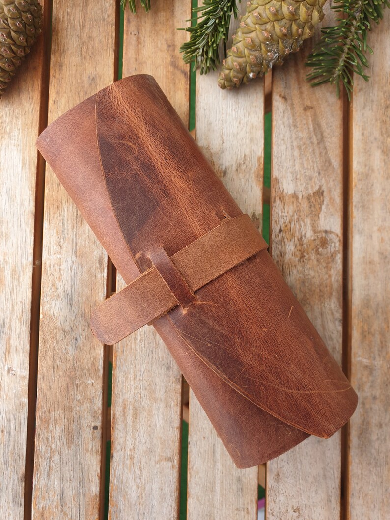 May include: A brown leather roll-up case with a strap closure. The case is made of distressed leather and has a rustic look.