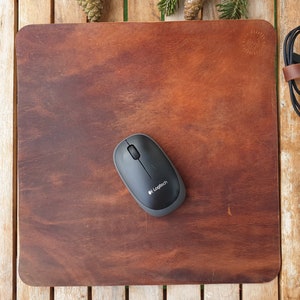 May include: A brown leather mouse pad with a black wireless mouse on top. The mouse pad has a small circular logo in the top right corner. The mouse is a Logitech brand.