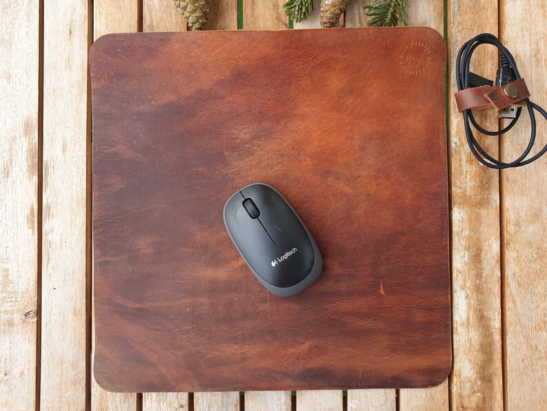 May include: A brown leather mouse pad with a black wireless mouse on top. The mouse pad has a small circular logo in the top right corner. The mouse is a Logitech brand.