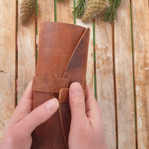 May include: A brown leather journal with a strap closure. The journal is rolled up and held in a person's hands.