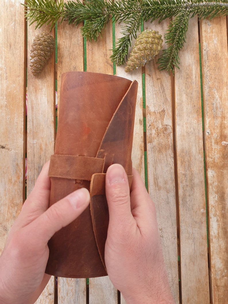 May include: A brown leather journal with a strap closure. The journal is rolled up and held in a person's hands.