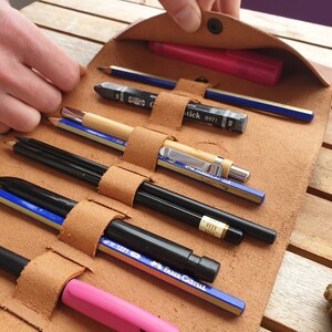 May include: A brown leather pencil roll with multiple slots for pencils, pens, and markers. The roll is open and the pencils are visible. The pencils include a black marker, a pink highlighter, a blue pencil, a wooden pencil, a black mechanical pencil, and a silver mechanical pencil.