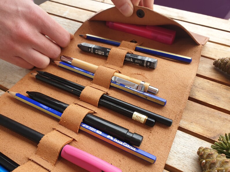 May include: A brown leather pencil roll with multiple slots for pencils, pens, and markers. The roll is open and the pencils are visible. The pencils include a black marker, a pink highlighter, a blue pencil, a wooden pencil, a black mechanical pencil, and a silver mechanical pencil.