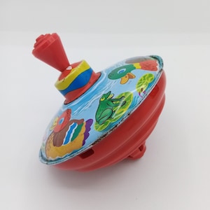 Könnte beinhalten: Ein rotes und blaues Kreisel mit einem Bild von Enten und Fröschen darauf. Der Kreisel ist aus Metall und hat einen kleinen Griff oben.