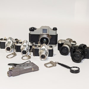 May include: A collection of miniature silver and black toy cameras, including a larger grey camera with a black lens. The cameras are all on a white surface.
