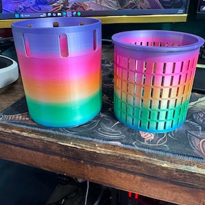 Könnte beinhalten: Zwei regenbogenfarbene, zylindrische Behälter. Der größere hat vertikale Schlitze und einen festen Boden. Der kleinere hat ein gitterartiges Muster aus rechteckigen Öffnungen. Beide sind 3D-gedruckt und farblich abgestuft, von Lila nach Grün.