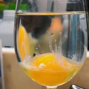 Puede incluir: Una copa de vino transparente llena de agua, que contiene una yema de huevo amarilla brillante y clara de huevo. La yema es redonda e intacta, mientras que la clara está parcialmente dispersa en el agua. La copa está sobre una superficie marrón claro.