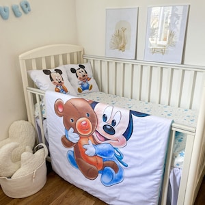 May include: White crib with a white and blue Mickey Mouse bedding set. The bedding features a large image of Mickey Mouse holding a brown teddy bear. The crib is decorated with two framed prints of a giraffe and a floral design.