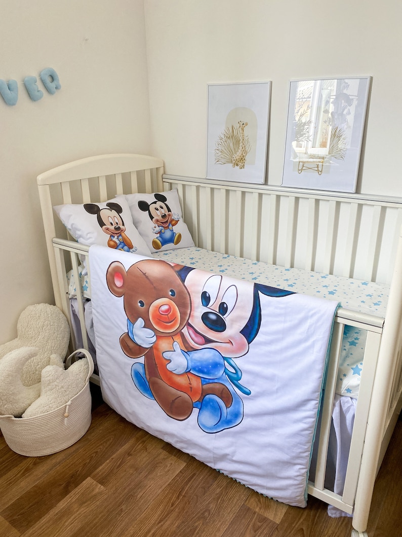 May include: White crib with a white and blue Mickey Mouse bedding set. The bedding features a large image of Mickey Mouse holding a brown teddy bear. The crib is decorated with two framed prints of a giraffe and a floral design.