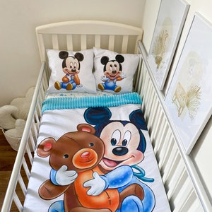 May include: A white crib bedding set with a blue and white Mickey Mouse print. The set includes a fitted sheet, a flat sheet, and two pillowcases. The fitted sheet features a large image of Mickey Mouse holding a brown teddy bear. The pillowcases feature a smaller image of Mickey Mouse in a blue and white outfit.