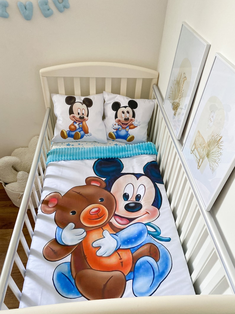 May include: A white crib bedding set with a blue and white Mickey Mouse print. The set includes a fitted sheet, a flat sheet, and two pillowcases. The fitted sheet features a large image of Mickey Mouse holding a brown teddy bear. The pillowcases feature a smaller image of Mickey Mouse in a blue and white outfit.