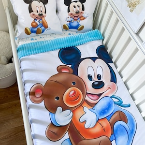 May include: A white crib bedding set with a blue and white star pattern. The set includes a fitted sheet, a crib blanket, and two pillowcases. The pillowcases feature a cartoon image of Mickey Mouse holding a teddy bear.