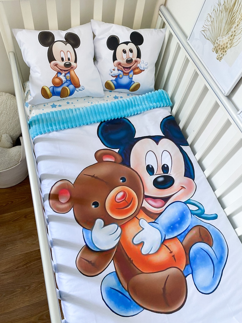 May include: A white crib bedding set with a blue and white star pattern. The set includes a fitted sheet, a crib blanket, and two pillowcases. The pillowcases feature a cartoon image of Mickey Mouse holding a teddy bear.