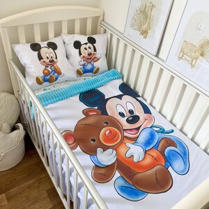 May include: A white crib with a white and blue Mickey Mouse bedding set. The bedding features a large image of Mickey Mouse holding a brown teddy bear. The crib also has two pillows with images of Mickey Mouse on them.