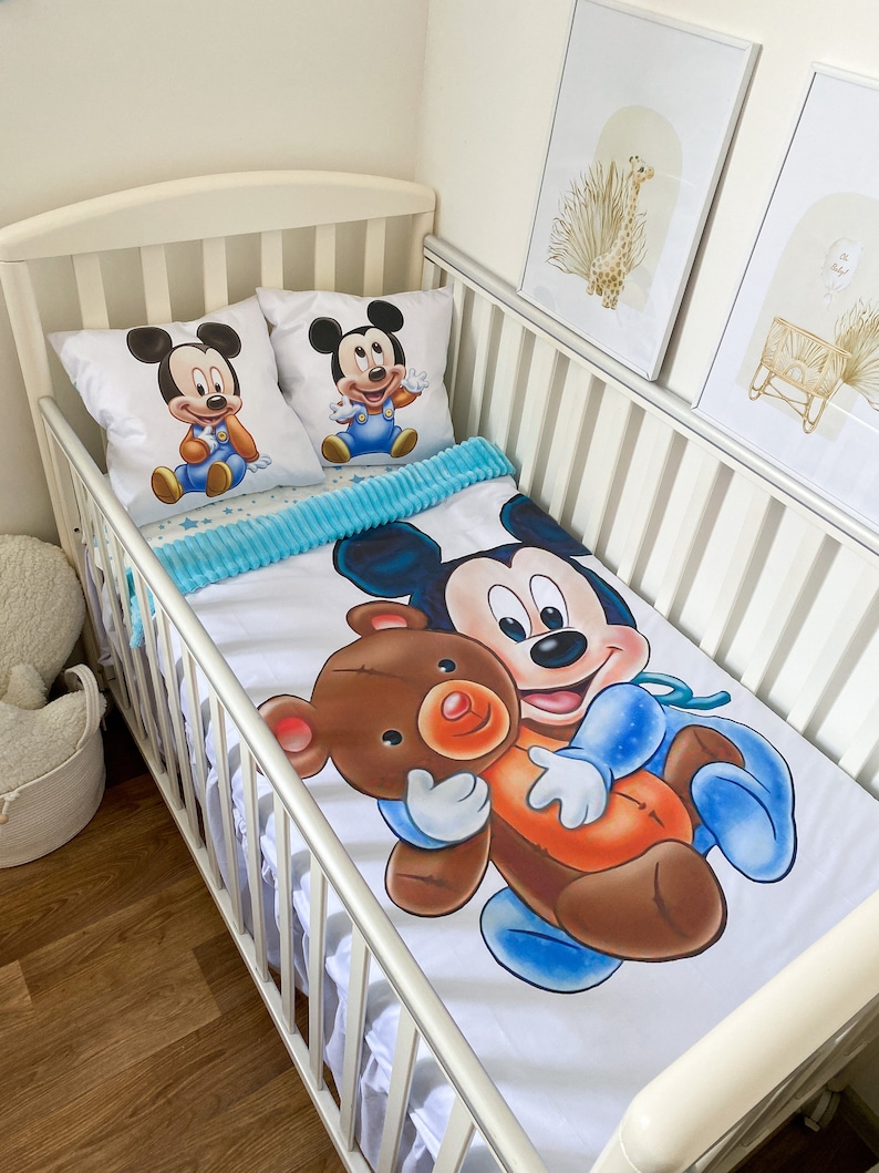 May include: A white crib with a white and blue Mickey Mouse bedding set. The bedding features a large image of Mickey Mouse holding a brown teddy bear. The crib also has two pillows with images of Mickey Mouse on them.