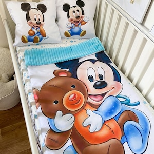 May include: A white crib bedding set with a blue and white star pattern. The set includes a fitted sheet, a crib blanket, and two pillowcases. The pillowcases feature a cartoon image of Mickey Mouse holding a teddy bear.