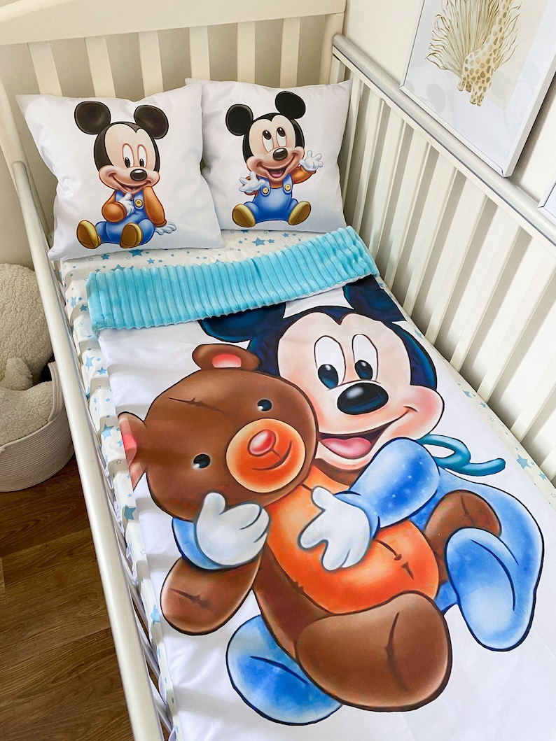 May include: A white crib bedding set with a blue and white star pattern. The set includes a fitted sheet, a crib blanket, and two pillowcases. The pillowcases feature a cartoon image of Mickey Mouse holding a teddy bear.