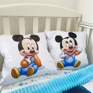 May include: Two white pillowcases with a blue and orange cartoon image of Mickey Mouse on each. The pillowcases are on a white crib with a blue fuzzy blanket.