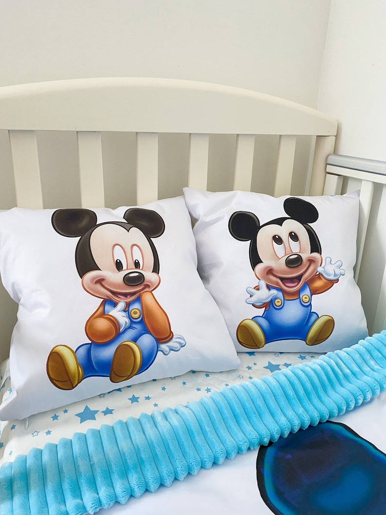 May include: Two white pillowcases with a blue and orange cartoon image of Mickey Mouse on each. The pillowcases are on a white crib with a blue fuzzy blanket.