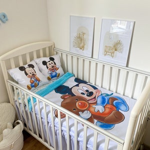May include: White crib with a white and blue Mickey Mouse bedding set. The bedding features a large Mickey Mouse holding a brown teddy bear. Two smaller Mickey Mouse pillows are on the crib.