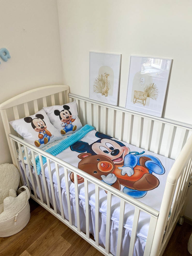 May include: White crib with a white and blue Mickey Mouse bedding set. The bedding features a large Mickey Mouse holding a brown teddy bear. Two smaller Mickey Mouse pillows are on the crib.