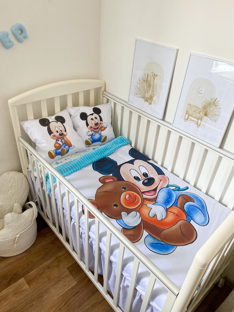 May include: White crib with a white and blue Mickey Mouse bedding set. The bedding features a large Mickey Mouse holding a brown teddy bear. The crib has a white ruffle around the bottom.