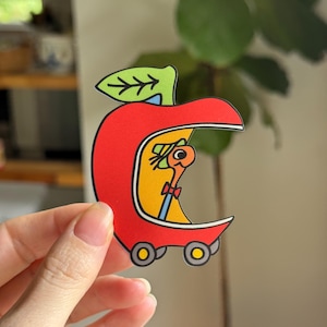 May include: A die-cut sticker shaped like a red apple car with a green leaf on top. Inside the apple, a tan worm wearing a green hat and a red bowtie is visible. The apple car has two grey wheels with yellow centers and a yellow interior.
