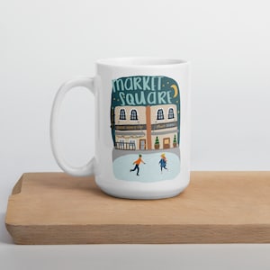 May include: White ceramic mug with a blue and white illustration of a building with the text "Market Square" and two people ice skating in front of it.