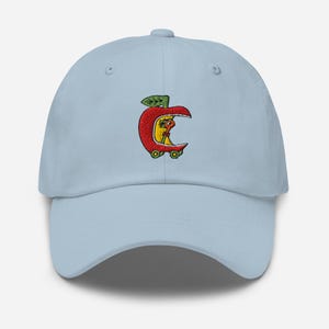 May include: Light blue baseball cap with an embroidered design of a red apple with a yellow interior and a green leaf. The apple has wheels and a small figure inside. The cap has a curved brim and an adjustable strap.