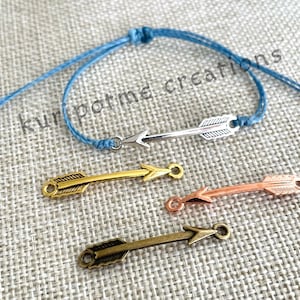 May include: A blue string bracelet with a silver arrow charm. There are also three arrow charms in gold, copper, and antique brass.
