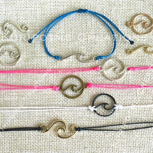 May include: Assortment of wave-themed bracelets with different colored cords and metal charms. The charms are in the shape of a wave, a circle with a wave inside, and a hook. The bracelets are made with a variety of colors, including blue, pink, black, and white.
