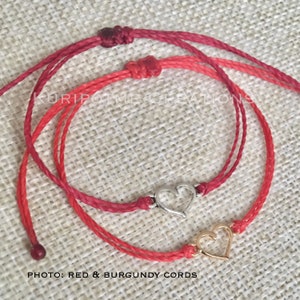 May include: Two friendship bracelets with heart charms. One bracelet is red and the other is burgundy. The heart charms are silver and gold.