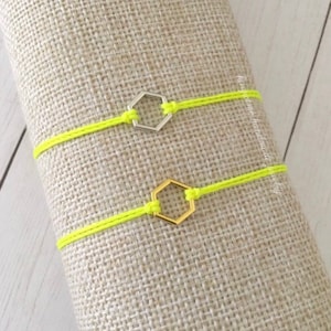 May include: Two neon yellow string bracelets. One features a silver hexagon charm, the other a gold hexagon charm. The bracelets are displayed on a textured, neutral-colored surface.