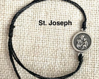 May include: A black cord bracelet featuring a round silver-toned metal charm with a relief image of St. Joseph holding the child Jesus. The charm is inscribed with the text St. Joseph. The bracelet has adjustable sliding knots at the ends.