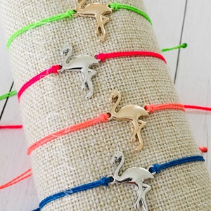 May include: Four colorful string bracelets with gold and silver flamingo charms. The bracelets are displayed on a beige fabric background.
