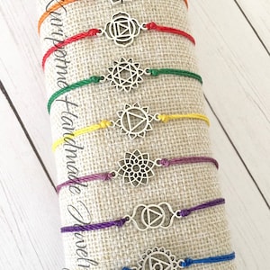 May include: Seven colorful string bracelets with silver charms depicting the seven chakras. The bracelets are arranged vertically on a beige fabric background. The text "Kumihimo Handmade Jewelry" is visible on the left side of the image.