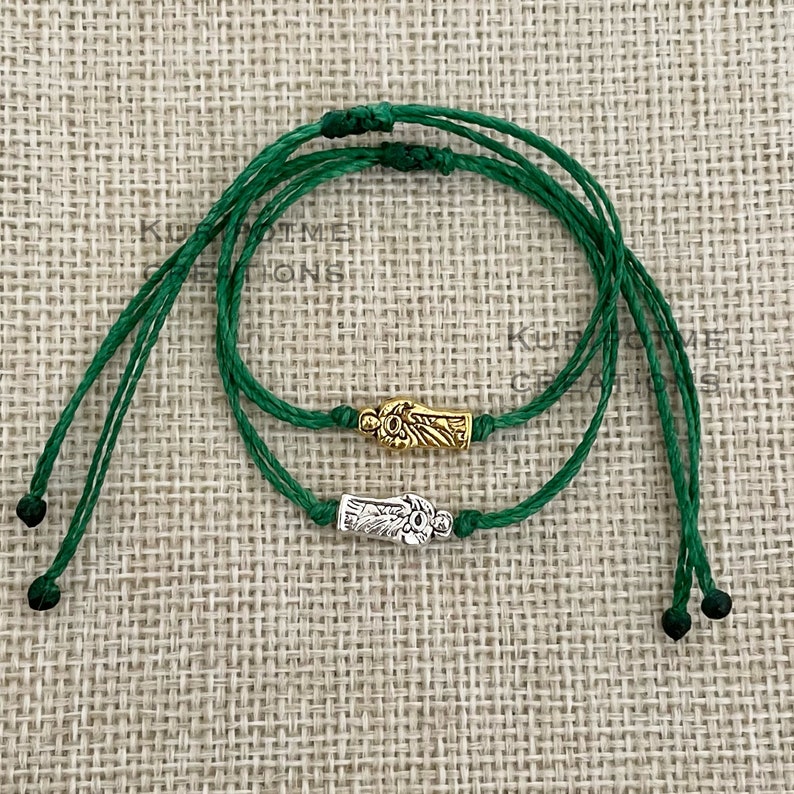 May include: Two adjustable green cord bracelets featuring small metal charms. One bracelet has a gold-toned charm depicting a figure, while the other has a silver-toned charm of the same design. Both bracelets have knotted ends and adjustable sliding closures.