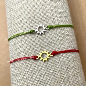 May include: Two string bracelets with a sun charm. The top bracelet has a silver sun charm and a green string. The bottom bracelet has a gold sun charm and a red string. Both bracelets have the text "Kuripotme Creations" on the string.