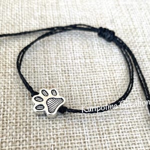 Puede incluir: Un brazalete negro ajustable con un dije de plata en forma de huella de pata. El brazalete está hecho de cordón delgado y tiene un diseño simple.