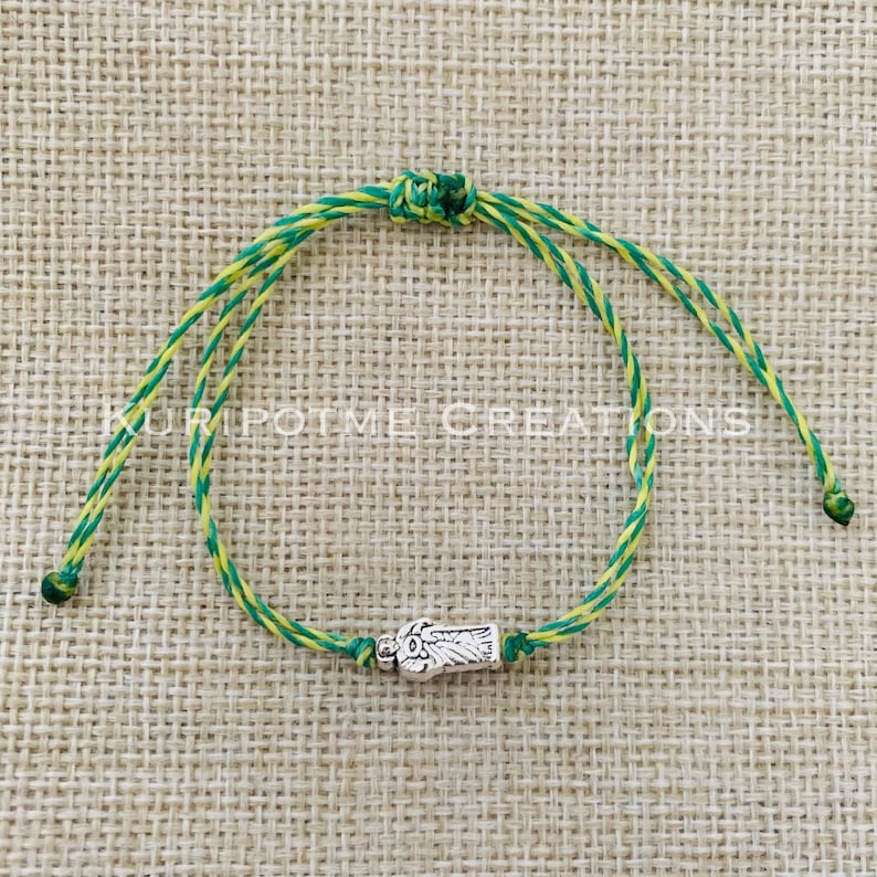 May include: A bracelet made of twisted yellow and green cord featuring a silver-toned metal charm shaped like a hand. The charm is centered on the cord, which includes a sliding knot mechanism. The text KURIPOTME CREATIONS is visible in white.