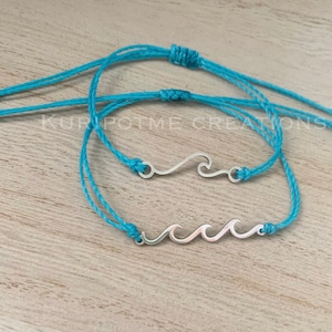 May include: Two turquoise blue friendship bracelets with silver wave charms. The bracelets are tied with a knot and have a small bead at each end.