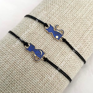 May include: Two black cord bracelets featuring gold-toned metal cat charms with blue enamel inlay. Each cat charm displays a crescent moon and star pattern on its body. The bracelets are thin, adjustable, and designed with a simple, minimalist aesthetic.
