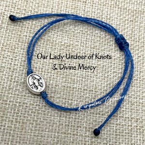 May include: Blue string bracelet with a silver charm featuring Our Lady Undoer of Knots & Divine Mercy. The bracelet is tied with a knot and has a small bead at each end.
