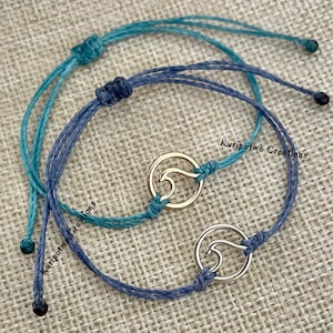 May include: Two adjustable bracelets with wave charms. One bracelet is teal, the other is blue. The wave charms are gold and silver. The text "Kuripotme Creations" is visible on the image.