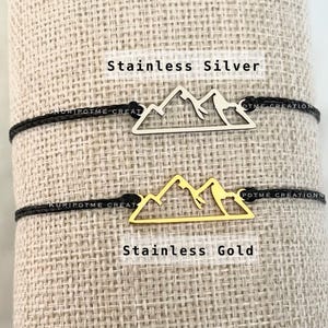 May include: Two black cord bracelets with mountain charm pendants. The top bracelet has a silver mountain charm, and the bottom bracelet has a gold mountain charm. The text "Stainless Silver" is above the silver charm, and the text "Stainless Gold" is below the gold charm.