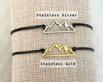May include: Two black cord bracelets with mountain charm pendants. The top bracelet has a silver mountain charm, and the bottom bracelet has a gold mountain charm. The text "Stainless Silver" is above the silver charm, and the text "Stainless Gold" is below the gold charm.