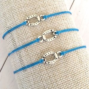 May include: Three bracelets featuring bright blue cord bands and silver-toned metal horseshoe charms. Each charm is secured to the cord with small loops. The text Kuripotme Creations is visible vertically along the left side of the image.