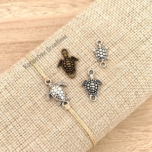 May include: A close-up of four silver turtle charms. Three of the charms are hanging from a beige string bracelet. The fourth charm is on a beige fabric surface. The charms have a detailed turtle design.