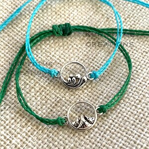 May include: Two friendship bracelets with a wave and mountain charm. The bracelets are made with turquoise blue and green cord.